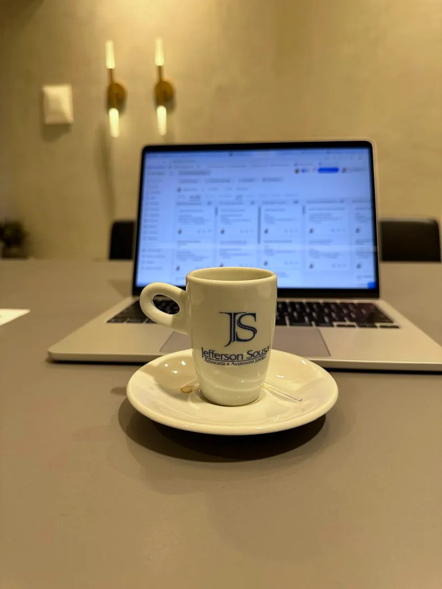 Xícara com a marca JS ao lado de um notebook sobre a mesa